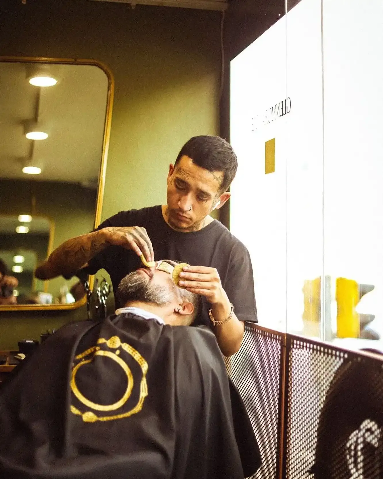Un barbero atiende el rostro de un cliente reclinado junto a la ventana del estudio.